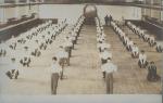 Real photo postcard, view of an activity in gymnasium, six columns of young men are arranged facing the camera, four of the six are squatting, two (in the middle) are standing with their hands on their shoulders, taken from vantage point of the balcony