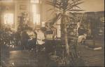 a real photo postcard, view of the school library, large plant in the foreground, in middleground to the left there are three tables with students looking at books, the background is a set of shelves to the right and a window and door to the left