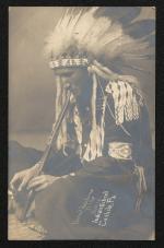 sepia-toned portrait of a young man sitting on the ground, he wears regalia and is lighting a pipe