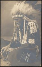 Daniel Hashorns, c.1907 sepia-toned portrait of a young man sitting on the ground, he wears regalia and is lighting a pipe