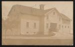 real photo postcard, sepia toned, a view of a two-storey building with a large door in the middle, driveway runs up to the door, three men stand around the door