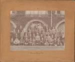 sepia toned photograph of 51 men sitting and standing in rows in front of ivy covered stonework