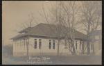 real photo postcard; view of the front of the printing office at the school (single story building with white walls and a dark gray roof), trees (without leaves) are in front of the building