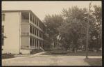 real photo postcard; view of the side and front of the girls' quarters (three story building with white walls and a porch for each level). Just out of view to the right would have been the band stand