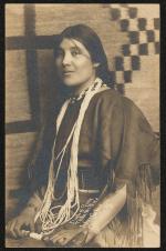real photo postcard, portrait of a young woman at the 3/4 angle. She sits in front of a rug or tapestry and wears regalia and jewelry